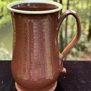 May include: A tall, cylindrical ceramic mug with a glossy, deep brown glaze and a smooth, light cream-colored rim. The mug features a vertical, slightly curved handle attached to the side and a raised, circular base, showcasing a simple, functional design.
