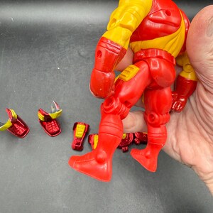 May include: A red and yellow action figure of a superhero, with detachable accessories. The figure is mostly red with yellow accents on the shoulders and upper arms. Several red and yellow accessories are scattered nearby.