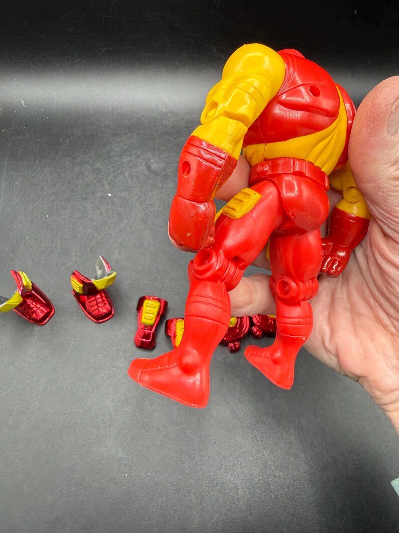 May include: A red and yellow action figure of a superhero, with detachable accessories. The figure is mostly red with yellow accents on the shoulders and upper arms. Several red and yellow accessories are scattered nearby.