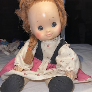 May include: A vintage doll with a painted face, rosy cheeks, and red hair in a braid. The doll wears a cream-colored dress with a pink skirt and black vest. The doll's black shoes are visible.