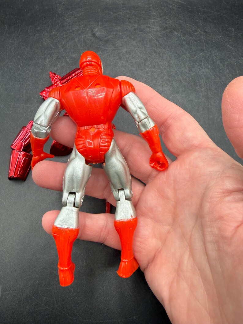 Puede incluir: Figura de acci&oacute;n roja y plateada de un superh&eacute;roe, vista desde atr&aacute;s. La figura tiene un casco, torso y piernas rojos, con brazos y parte inferior de las piernas plateados. La figura es articulada y tiene un accesorio rojo met&aacute;lico.