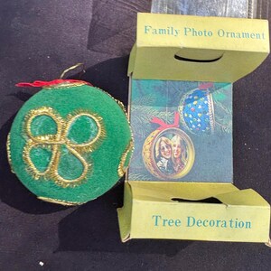 May include: A green Christmas ornament with gold trim and a red ribbon. The ornament is next to a box labeled "Family Photo Ornament" and "Tree Decoration". The box shows a photo ornament with two women.