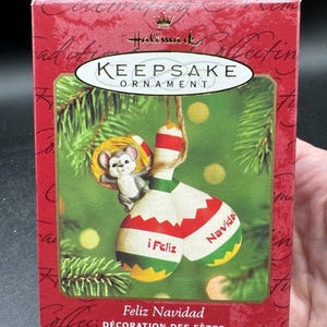 May include: A Hallmark Keepsake Christmas ornament in a red box. The ornament features a mouse holding onto a red and white striped ornament with the words "i Feliz" and "Navidad". The box reads "Feliz Navidad" and "Décoration des Fêtes".