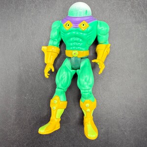 Op de afbeelding: Een groene actiefiguur met een heldere, afgeronde kop en paarse accenten. De figuur heeft gele handschoenen, laarzen en een riem. De figuur staat rechtop met de armen langs de zijden.