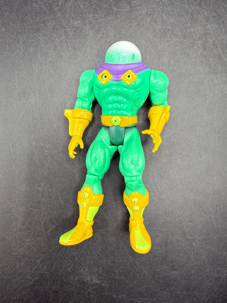 Op de afbeelding: Een groene actiefiguur met een heldere, afgeronde kop en paarse accenten. De figuur heeft gele handschoenen, laarzen en een riem. De figuur staat rechtop met de armen langs de zijden.