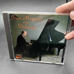 May include: A compact disc case featuring a cover with a photograph of a pianist playing a black grand piano. The text reads Ivan Moravec plays Debussy, Images (Complete), Estampes, and Des pas sur la neige, with an MMG logo in the corner.