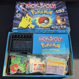 May include: A Monopoly Pokémon Collector's Edition board game box and open base. The box lid features Pikachu, Charmander, and a Poké Ball with the text Monopoly Pokémon Gotta catch 'em all!. The open box contains game pieces in plastic bags.