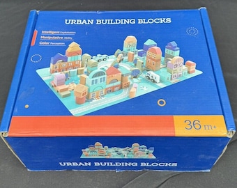 Könnte beinhalten: Eine blaue Box mit einem roten Streifen und dem Text "URBAN BUILDING BLOCKS" auf der Vorderseite. Die Box enthält einen Satz bunter Holzbausteine, mit denen eine Stadtlandschaft gebaut werden kann. Die Bausteine sind in einem Stadtlayout auf einer blauen und grünen Matte angeordnet. Die Box trägt auch den Text "36 m+" auf der Vorderseite.