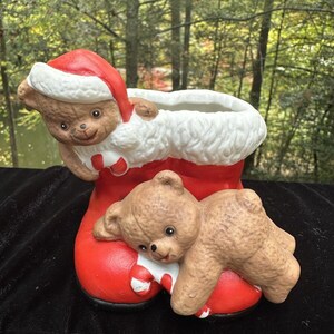 May include: A red and white ceramic Christmas boot planter with two brown teddy bears. The boot has a white fur trim and a candy cane design. One bear is peeking out of the boot and the other is lying on the boot.