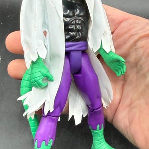 K&ouml;nnte beinhalten: Eine gr&uuml;ne, echsenartige Actionfigur mit einem wei&szlig;en Laborkittel, einer schwarzen Brust und einer lila Hose. Die Figur hat einen reptilienartigen Kopf mit roten Augen und einen wei&szlig;en Laborkittel mit gezackten Kanten. Die F&uuml;&szlig;e und H&auml;nde der Figur sind gr&uuml;n.