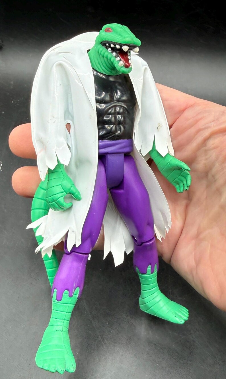 K&ouml;nnte beinhalten: Eine gr&uuml;ne, echsenartige Actionfigur mit einem wei&szlig;en Laborkittel, einer schwarzen Brust und einer lila Hose. Die Figur hat einen reptilienartigen Kopf mit roten Augen und einen wei&szlig;en Laborkittel mit gezackten Kanten. Die F&uuml;&szlig;e und H&auml;nde der Figur sind gr&uuml;n.