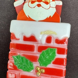 May include: A vintage Christmas decoration featuring Santa Claus emerging from a red brick chimney. The chimney has white frosting and green holly accents. Santa is wearing a red suit and hat with a white beard.
