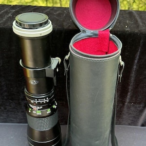 May include: A black camera lens with a silver ring and a black leather carrying case. The lens has a depth of field scale and a focus ring.