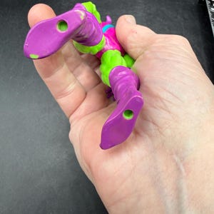 May include: A purple and green action figure of a comic book character. The figure has purple boots, legs, and arms, with green accents on the torso and head. The figure is held in a hand.