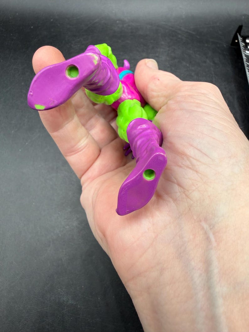 May include: A purple and green action figure of a comic book character. The figure has purple boots, legs, and arms, with green accents on the torso and head. The figure is held in a hand.