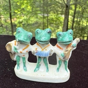 Puede incluir: Figura de cerámica que representa a tres ranas verdes en una banda. Cada rana viste una camisa blanca con detalles naranjas y dorados, y toca un instrumento diferente. La base es blanca.