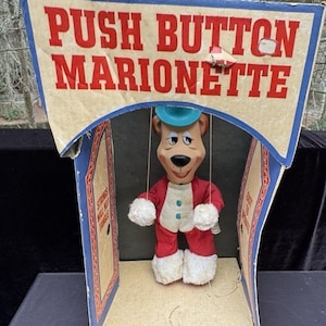 May include: Vintage push button marionette toy in its original box. The marionette is a bear wearing a red and white outfit with a blue hat. The box is labeled "Push Button Marionette".