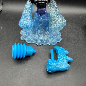 May include: A vintage action figure of a muscular man with a blonde head and black torso, blue water-like arms and base. Includes two blue accessories. The figure is a collectible toy.