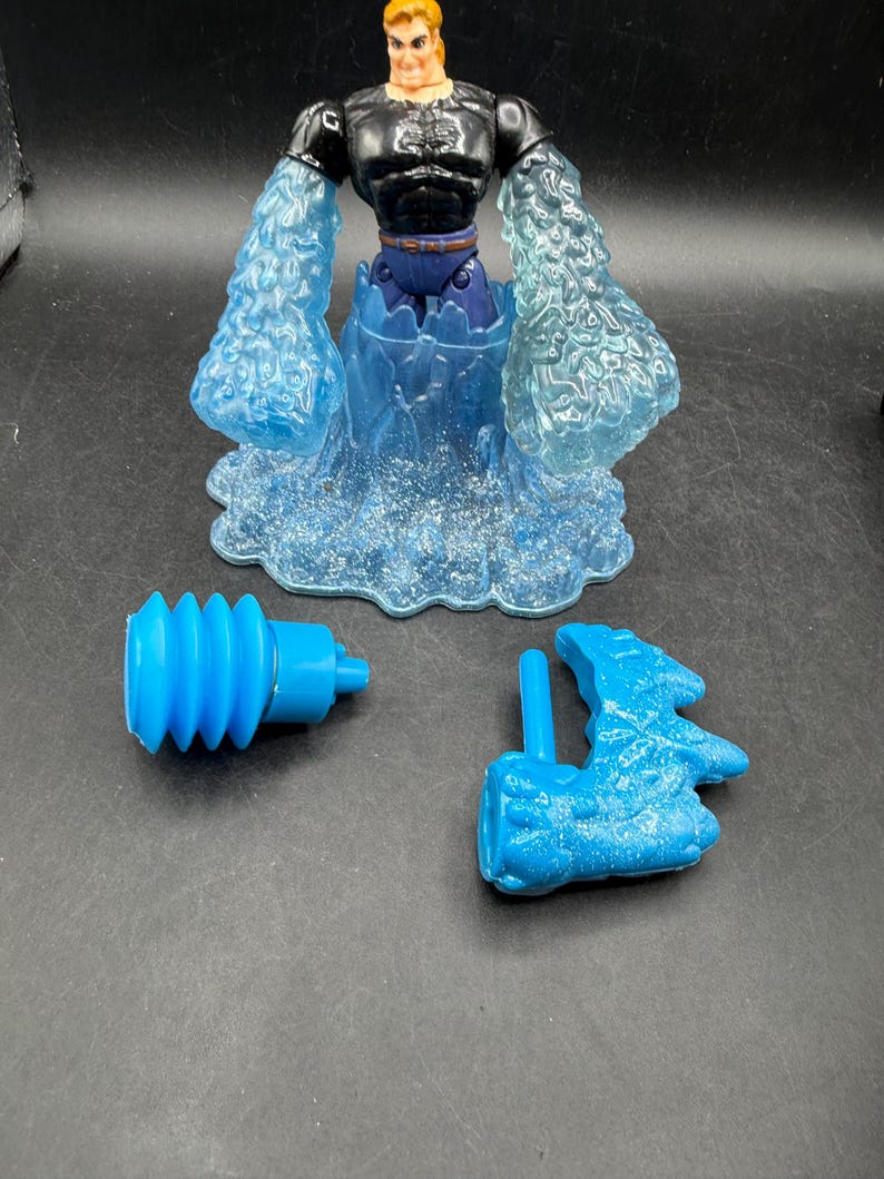 May include: A vintage action figure of a muscular man with a blonde head and black torso, blue water-like arms and base. Includes two blue accessories. The figure is a collectible toy.