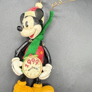 May include: A Mickey Mouse Christmas ornament featuring a character wearing a red and white Santa hat and a green felt scarf. The figure holds a round clock face with the year 1999 printed in red. It includes a gold wire hanger with beads.