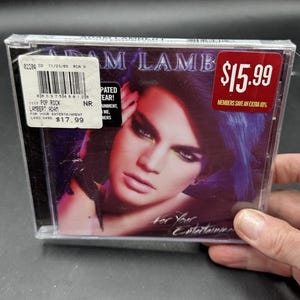 May include: A CD case featuring Adam Lambert's album "For Your Entertainment." The cover art showcases a close-up of Lambert with dramatic makeup and blue hair. The CD case has a price sticker with the price $15.99.