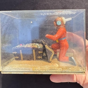 May include: A red and white plastic toy diver figurine kneeling next to a black treasure chest with gold coins inside. The figurine is in a clear plastic display case with a sandy base.