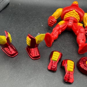 May include: A red and yellow Iron Man action figure with detachable parts. The figure is in a dynamic pose, with separate pieces including boots, arm pieces, and a chest plate. The figure's design features a metallic red finish with yellow accents.