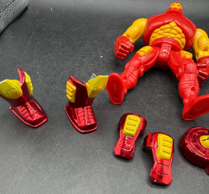 May include: A red and yellow Iron Man action figure with detachable parts. The figure is in a dynamic pose, with separate pieces including boots, arm pieces, and a chest plate. The figure's design features a metallic red finish with yellow accents.