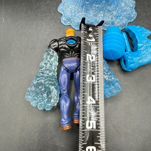 May include: A blue and black action figure with translucent blue ice-like arms, standing approximately 5 inches tall. The figure has a muscular build, blue pants, and brown boots. Additional blue plastic accessories are also present.