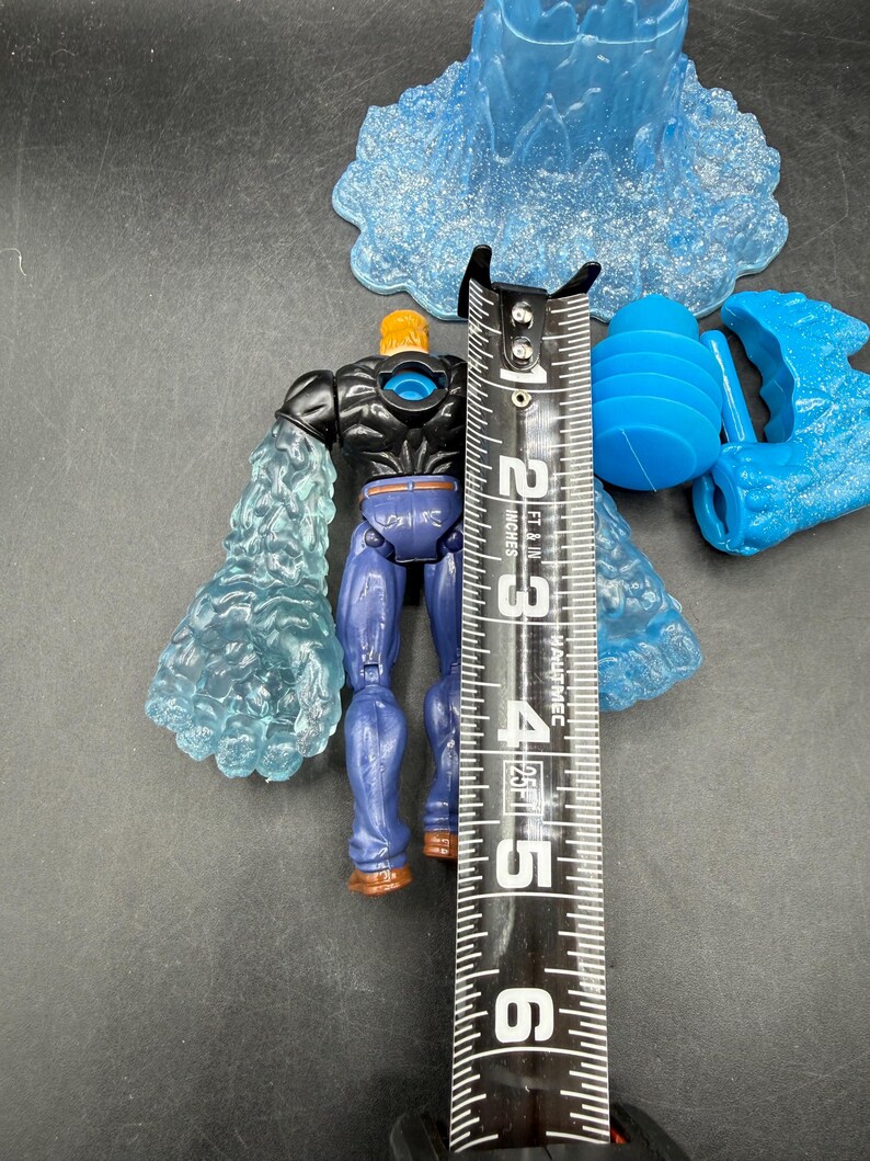 May include: A blue and black action figure with translucent blue ice-like arms, standing approximately 5 inches tall. The figure has a muscular build, blue pants, and brown boots. Additional blue plastic accessories are also present.