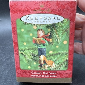Könnte beinhalten: Eine Hallmark Keepsake Ornament Schachtel mit dem Titel Caroler's Best Friend. Die rote Verpackung zeigt eine Illustration eines Kindes in grünem Mantel und rotem Schal, das aus einem Buch singt, neben einem braun-weißen Hund vor Tannen.