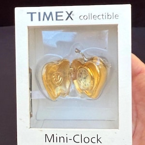 Może przedstawiać: Złoty, kolekcjonerski mini-zegar Timex w kształcie jabłka, prezentowany w białym pudełku. Tarcza zegara jest widoczna wewnątrz otwartego jabłka. Pudełko ma logo Timex oraz napisy "collectible" i "Mini-Clock".
