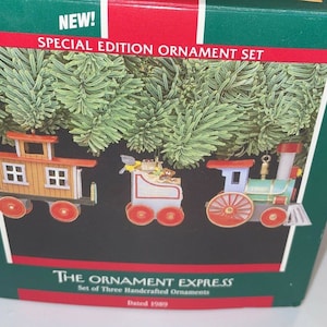 May include: A set of three vintage 1989 Christmas ornaments depicting a train. The ornaments are hand-painted and feature a red caboose, a blue and white passenger car, and a green engine. The box reads "The Ornament Express, Set of Three Handcrafted Ornaments, Dated 1989."