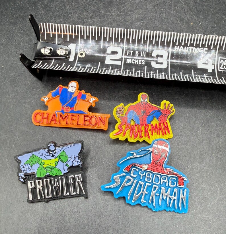 Może przedstawiać: Cztery kolorowe przypinki o tematyce superbohaterskiej. Przypinki przedstawiają postacie takie jak Spider-Man, The Prowler i Chameleon. Przypinki Spider-Man są czerwone i ż&oacute;łte, a The Prowler jest zielony i czarny. Przypinka Chameleon jest pomarańczowa i niebieska. W tle znajduje się linijka.