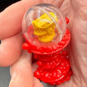 K&ouml;nnte beinhalten: Ein kleines, rotes Plastikspielzeug mit einer transparenten, kugelf&ouml;rmigen Kuppel, die ein gelbes Objekt umschlie&szlig;t. Der rote Sockel hat ein strukturiertes, geschichtetes Design. Das Spielzeug wird in der Hand einer Person gehalten.