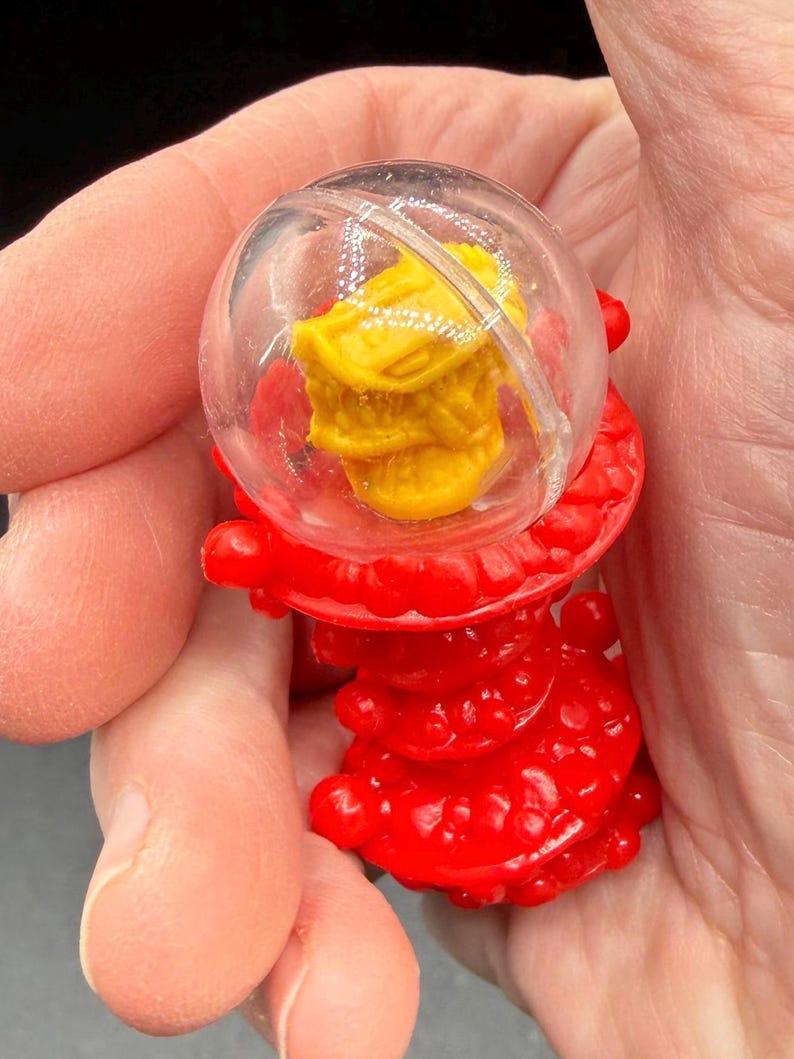 K&ouml;nnte beinhalten: Ein kleines, rotes Plastikspielzeug mit einer transparenten, kugelf&ouml;rmigen Kuppel, die ein gelbes Objekt umschlie&szlig;t. Der rote Sockel hat ein strukturiertes, geschichtetes Design. Das Spielzeug wird in der Hand einer Person gehalten.