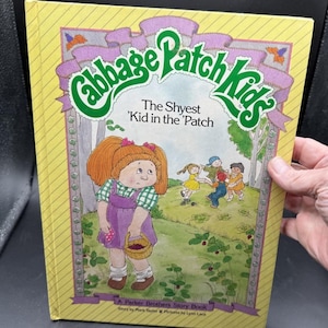 May include: A yellow hardcover storybook titled Cabbage Patch Kids: The Shyest 'Kid in the 'Patch. The cover features a central illustration of a character with orange pigtails holding a basket, with other children playing in a green field.