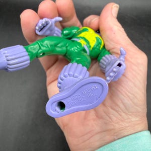May include: A vintage superhero action figure with a green body, yellow detailing, and purple limbs. The figure is held in a hand, displaying its design and the text "&copy;1995 MARVEL TOY BIZ INC." on the foot.