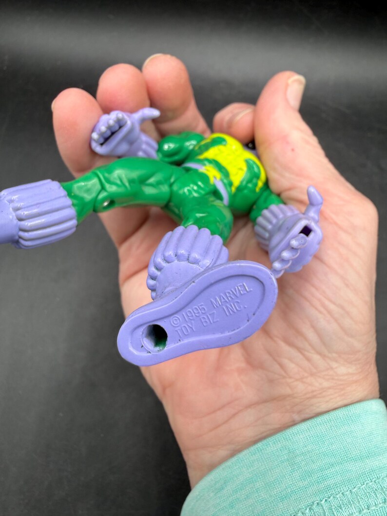 May include: A vintage superhero action figure with a green body, yellow detailing, and purple limbs. The figure is held in a hand, displaying its design and the text "&copy;1995 MARVEL TOY BIZ INC." on the foot.
