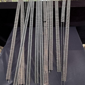 Könnte beinhalten: Eine Sammlung von Modellbahngleisen in verschiedenen Längen und Ausführungen. Die Gleise bestehen aus silberfarbenem Metall und braunen Holzschwellen. Die Gleise sind auf einer dunklen Oberfläche angeordnet, mit einem natürlichen Hintergrund.