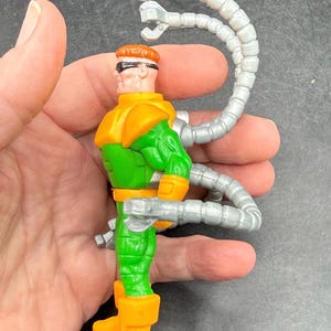 Puede incluir: Una figura de acci&oacute;n del Doctor Octopus. La figura es principalmente verde y naranja, con brazos mec&aacute;nicos plateados que se extienden desde su espalda. La figura mide aproximadamente 10 cm de altura.