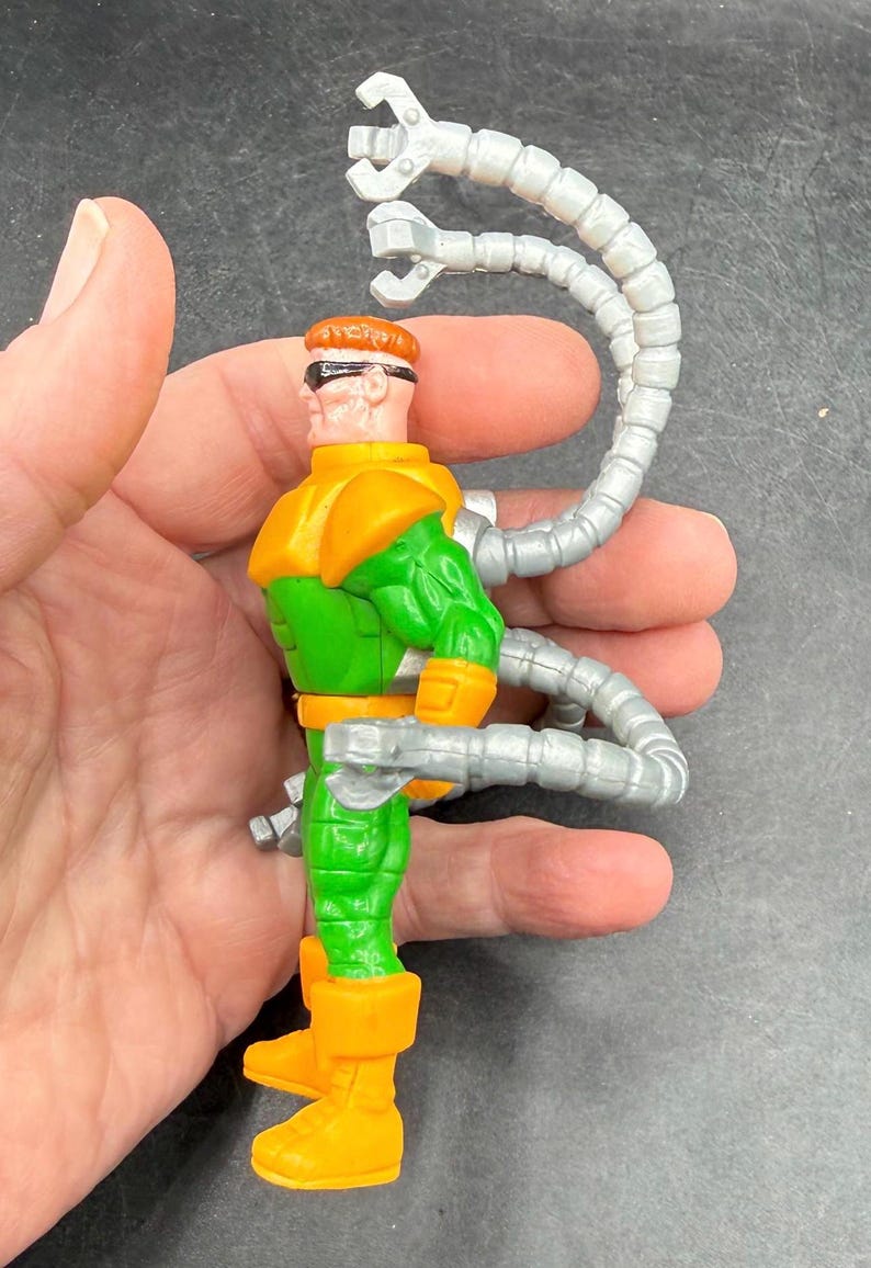 Puede incluir: Una figura de acci&oacute;n del Doctor Octopus. La figura es principalmente verde y naranja, con brazos mec&aacute;nicos plateados que se extienden desde su espalda. La figura mide aproximadamente 10 cm de altura.