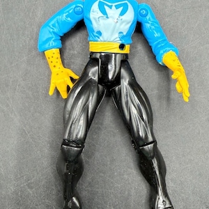 May include: Action figure of a character in a blue and white top with a yellow belt and gloves, and black pants. The figure has a stern expression and is posed with arms outstretched. The figure is a collectible toy.