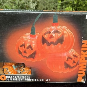 Op de afbeelding: Een doos met 20 superheldere pompoenlampjes voor binnen of buiten. De doos heeft een afbeelding van vier oranje pompoenen met pompoengezichten. De tekst op de doos luidt "20 Superbright Pumpkin Light Set".