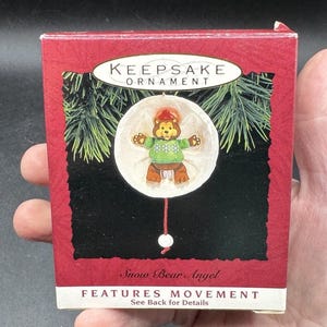 Może przedstawiać: Czerwone pudełko z ozdobą świąteczną. Ozdoba przedstawia misia w zielonym swetrze i czerwonej czapce. Na pudełku widnieje napis "Keepsake Ornament" i "Snow Bear Angel".