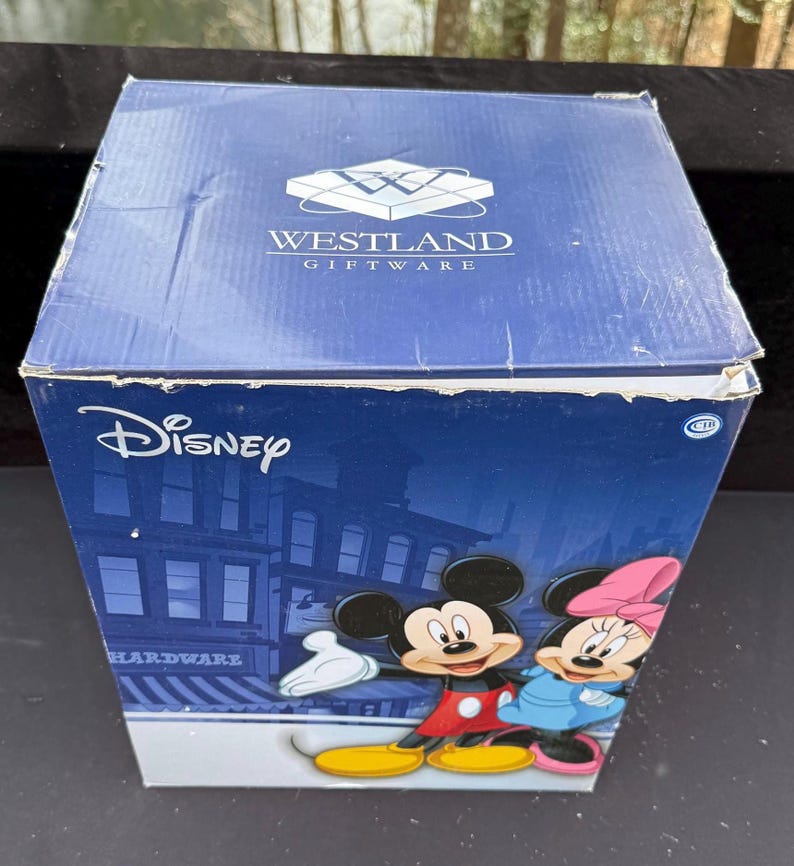 May include: A blue Westland Giftware box featuring Disney's Mickey Mouse and Minnie Mouse. The box has a cityscape background and the Disney logo. The box is a square shape and is made of cardboard.