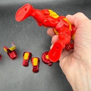 May include: A red and yellow Iron Man action figure with detachable armor pieces. The figure is held by a hand, showcasing its articulated design and vibrant colors. Several red and yellow armor components are scattered nearby.