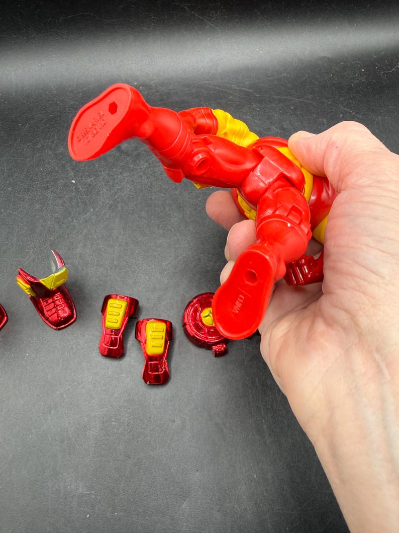 May include: A red and yellow Iron Man action figure with detachable armor pieces. The figure is held by a hand, showcasing its articulated design and vibrant colors. Several red and yellow armor components are scattered nearby.