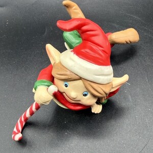 May include: A ceramic Christmas elf figurine with a red hat, green collar, and a candy cane. The elf has a smiling face with blue eyes and brown hair. The figurine is a festive holiday decoration.