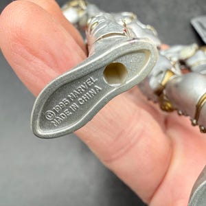 May include: A silver-colored action figure with gold accents. The figure's foot is visible, with the text "&copy;1995 MARVEL MADE IN CHINA" imprinted on the sole. The figure is held in a hand.